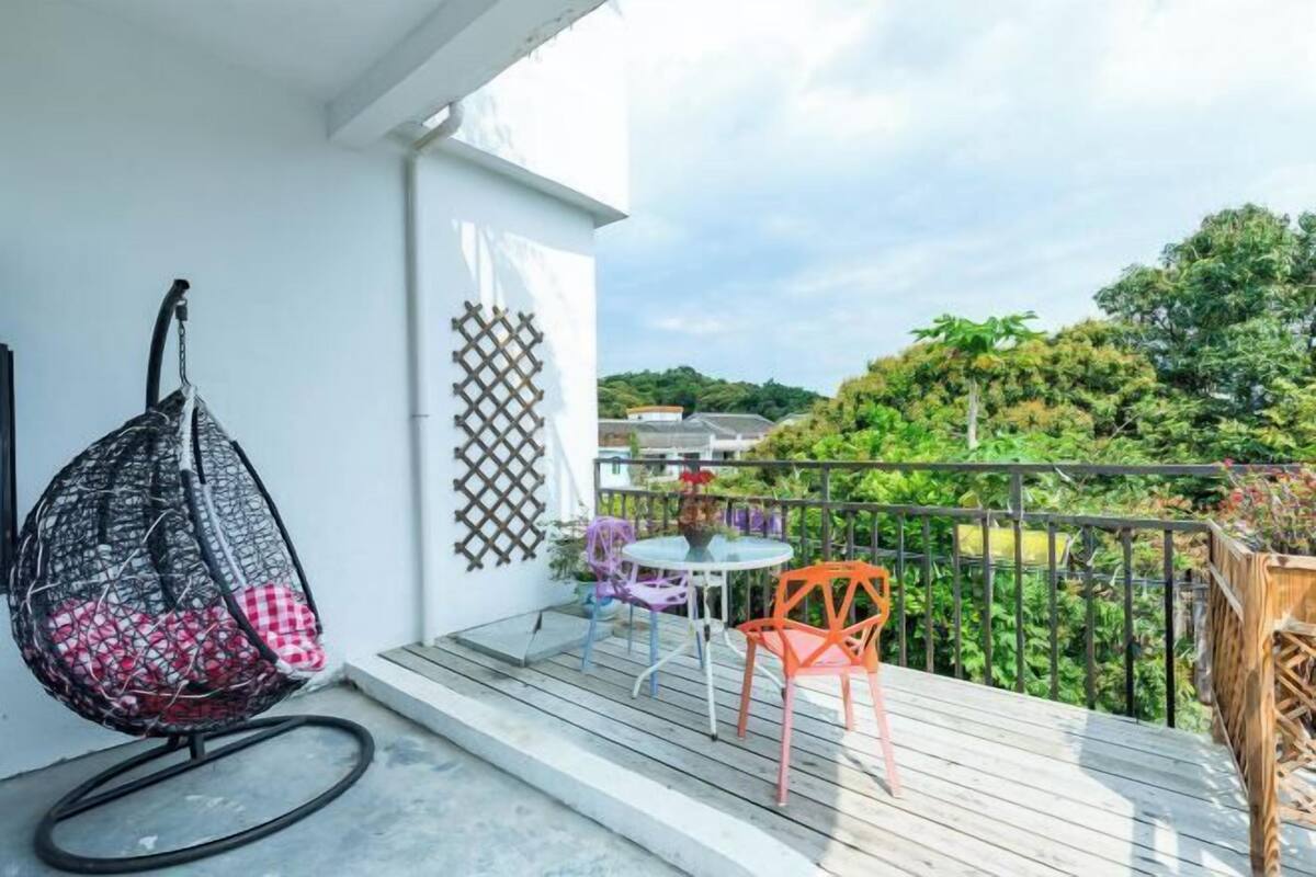 A balcony with a hanging chair, a small table with chairs, and a view of greenery and buildings.