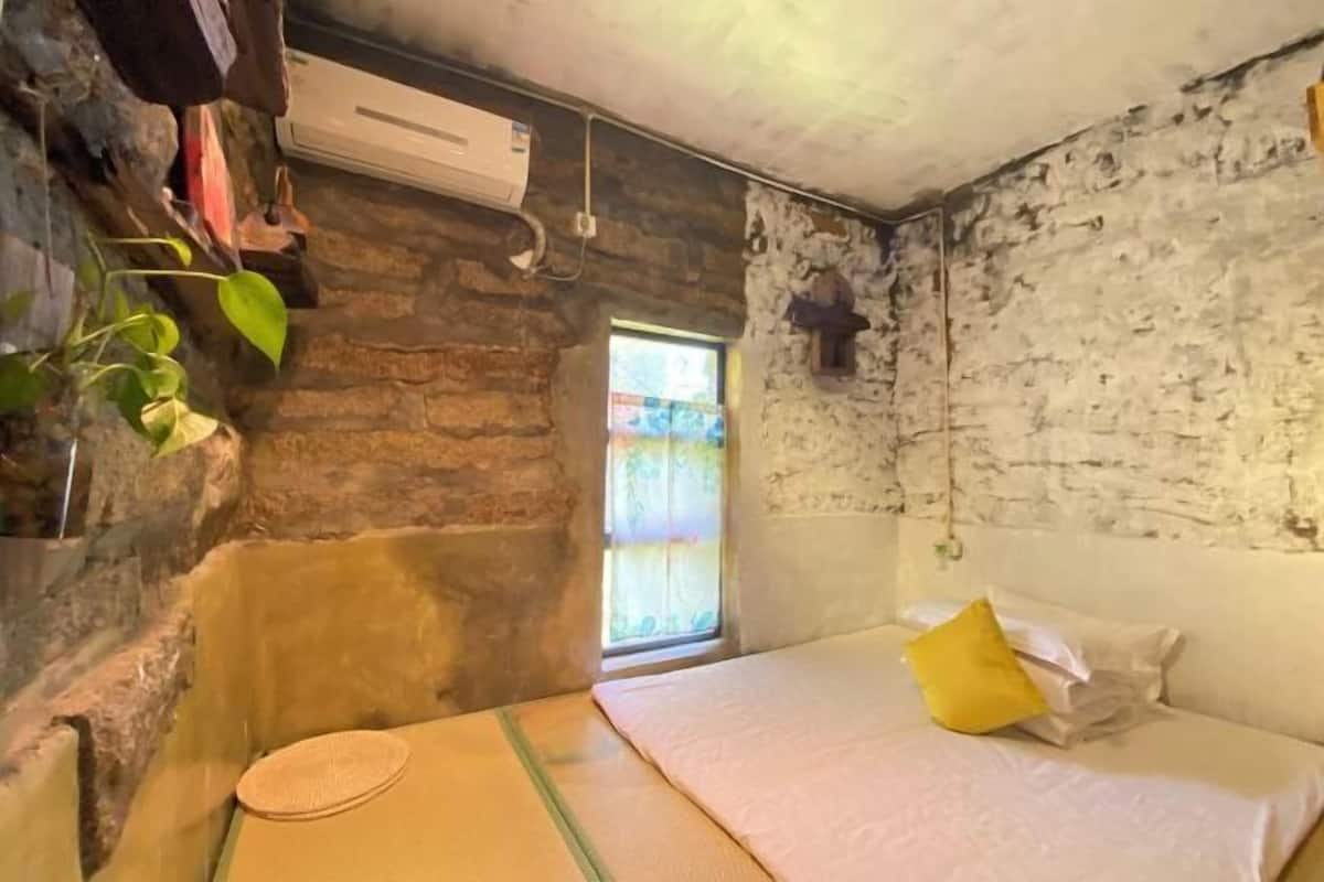 A room with a stone wall, a bed with a yellow pillow, a window with a view, and an air conditioning unit.