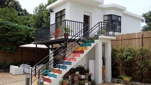Una casa moderna con una escalera que conduce a un balcón, rodeada de plantas en macetas y una terraza de madera.