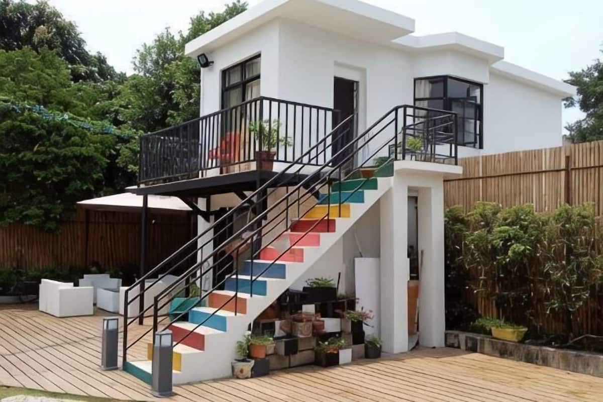 A modern house with a staircase leading to a balcony, surrounded by potted plants and a wooden deck.