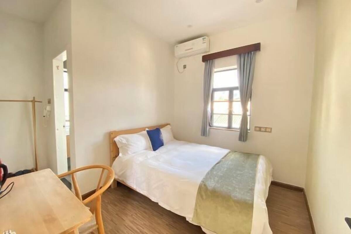 A single bed with a wooden frame, a table with a chair, a window with curtains, and an air conditioning unit on the wall.