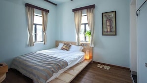 A bedroom with a bed, two windows with curtains, a bedside table with a lamp, and a wall-mounted artwork.