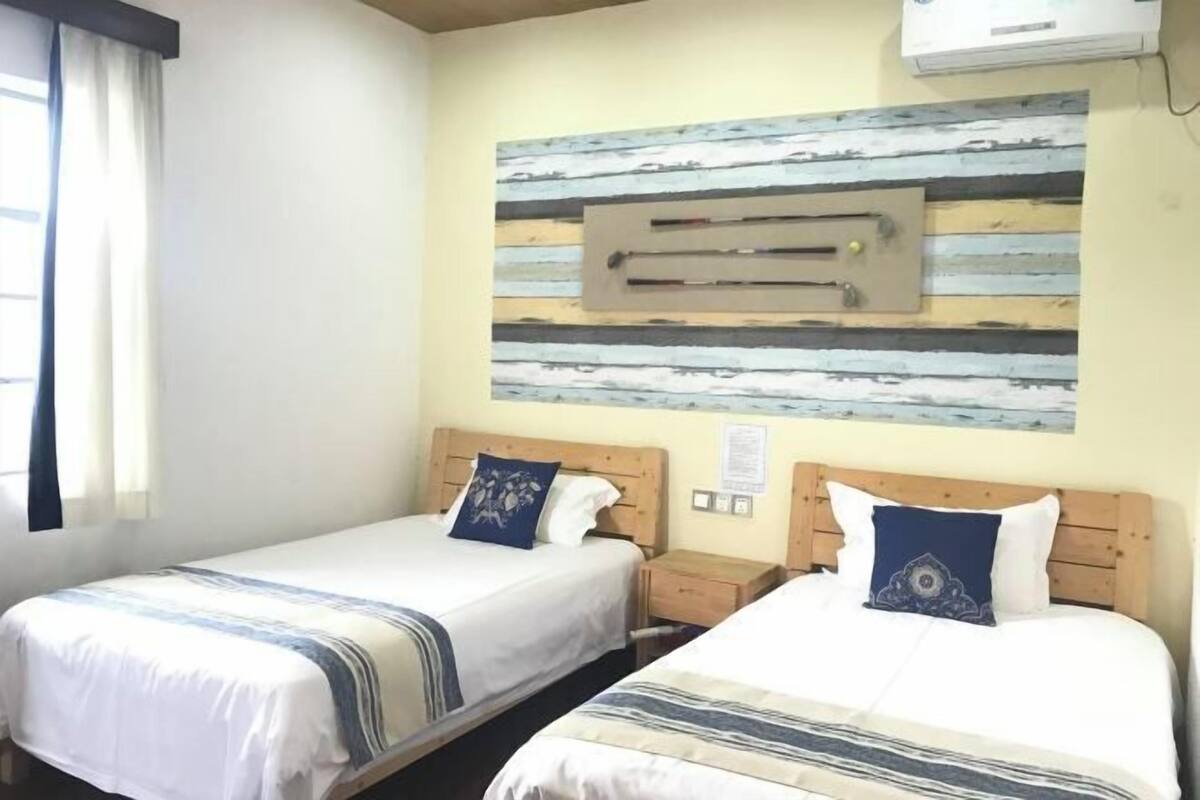 A hotel room with two beds, a wooden headboard, a wall-mounted air conditioning unit, and a window with curtains.