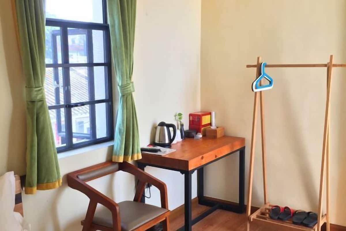 A room with a wooden desk, a chair, a window with green curtains, a wooden shoe rack, and a small table with a kettle and a box.