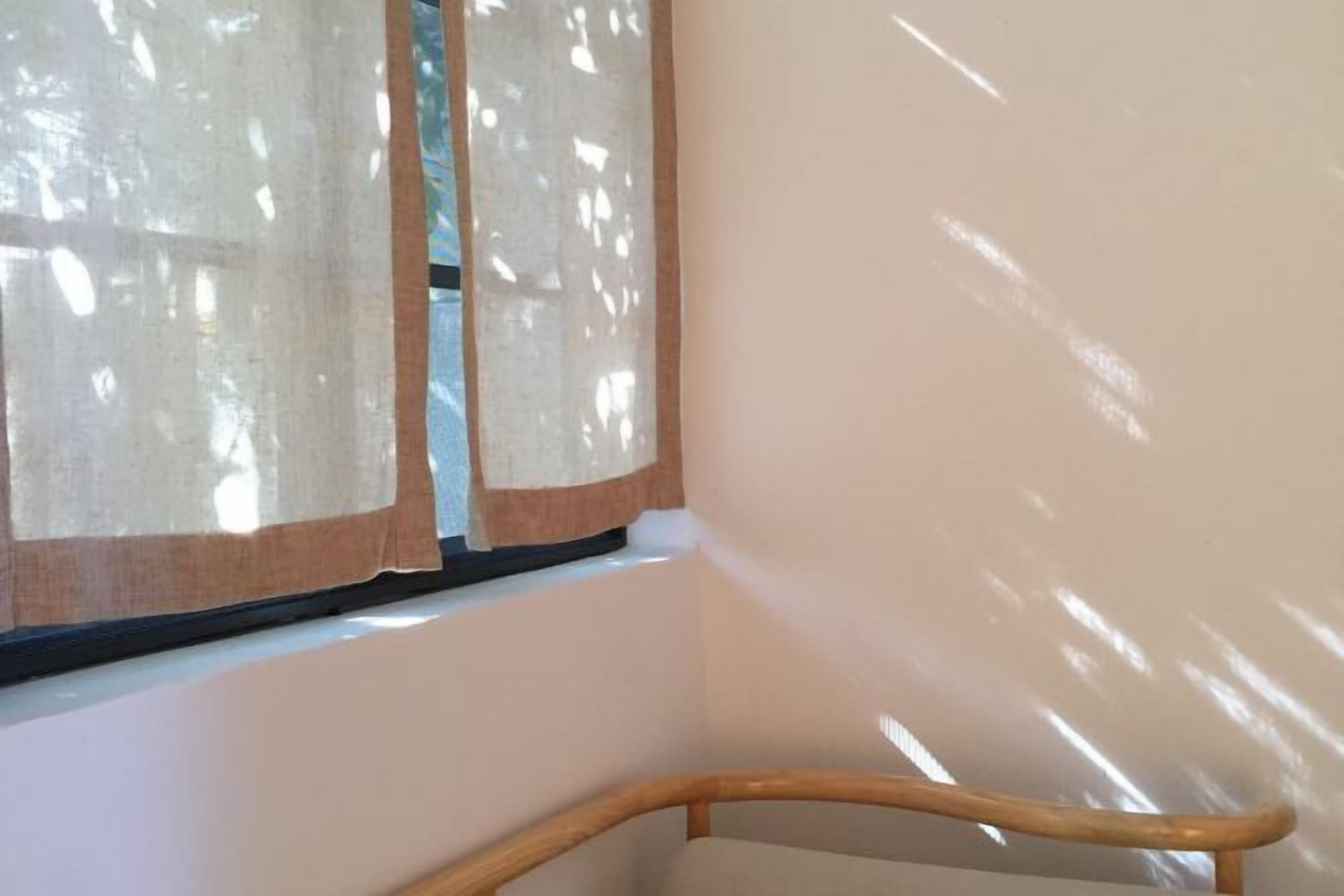 A corner of a room with a wooden chair, a window with sheer curtains, and a plain wall.