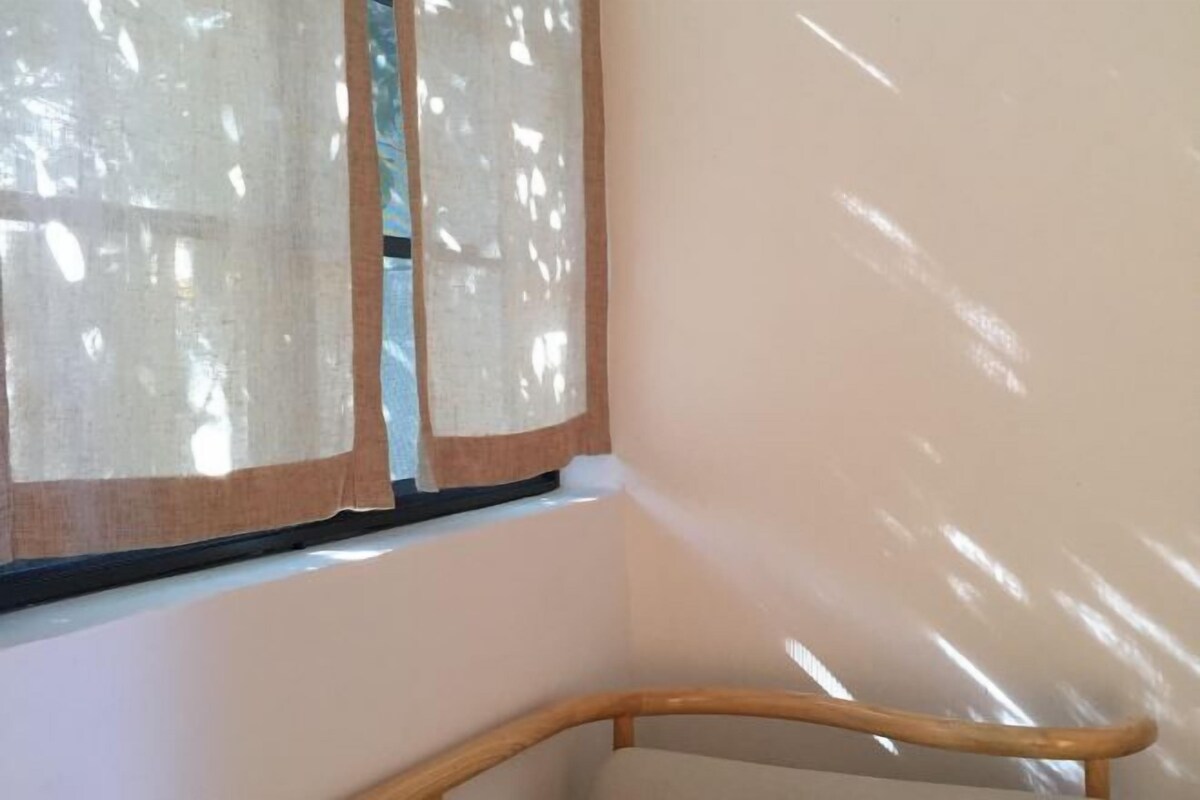 Una esquina de una habitación con una silla de madera, una ventana con cortinas traslúcidas y una pared lisa.