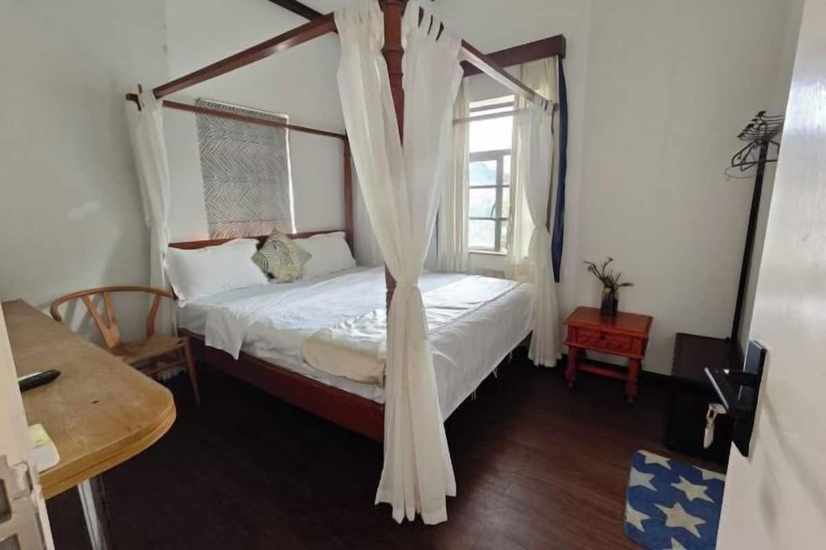 A room with a four-poster bed, a small table, a chair, a bedside table, and a window with curtains.