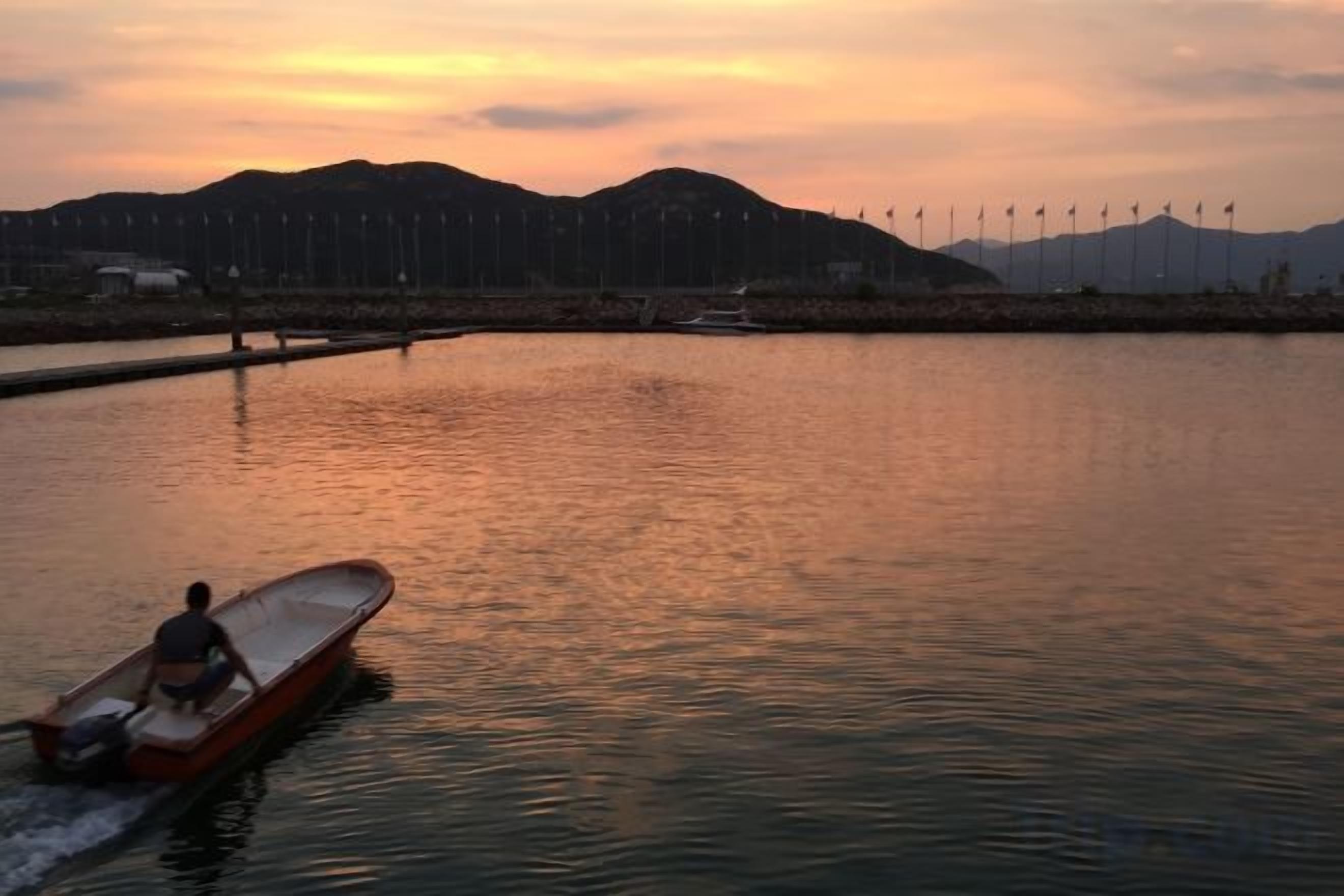 Un río tranquilo con una embarcación que se acerca, una orilla lejana con aerogeneradores y un atardecer.