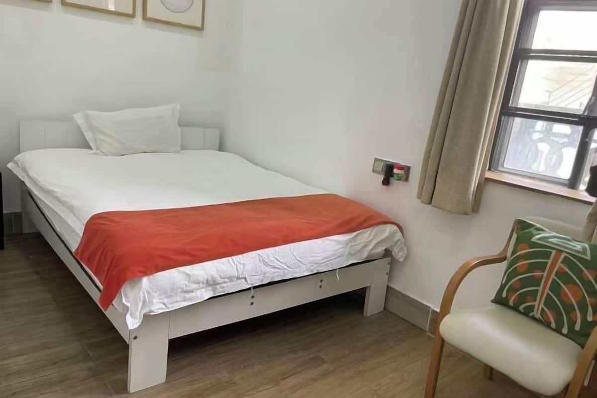 A single bed with a white and orange bedspread, a chair with a patterned cushion, and a window with curtains.