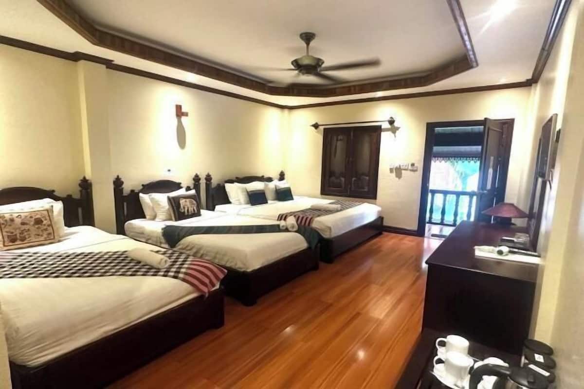 A hotel room with two beds, wooden flooring, a ceiling fan, a desk with a lamp, and a view of the outside.