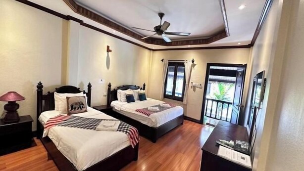 A hotel room with two beds, a ceiling fan, a nightstand with a lamp, a small table with items, and a view of the outdoors through a door. - Y Not Lao Villa (Luang Prabang)
