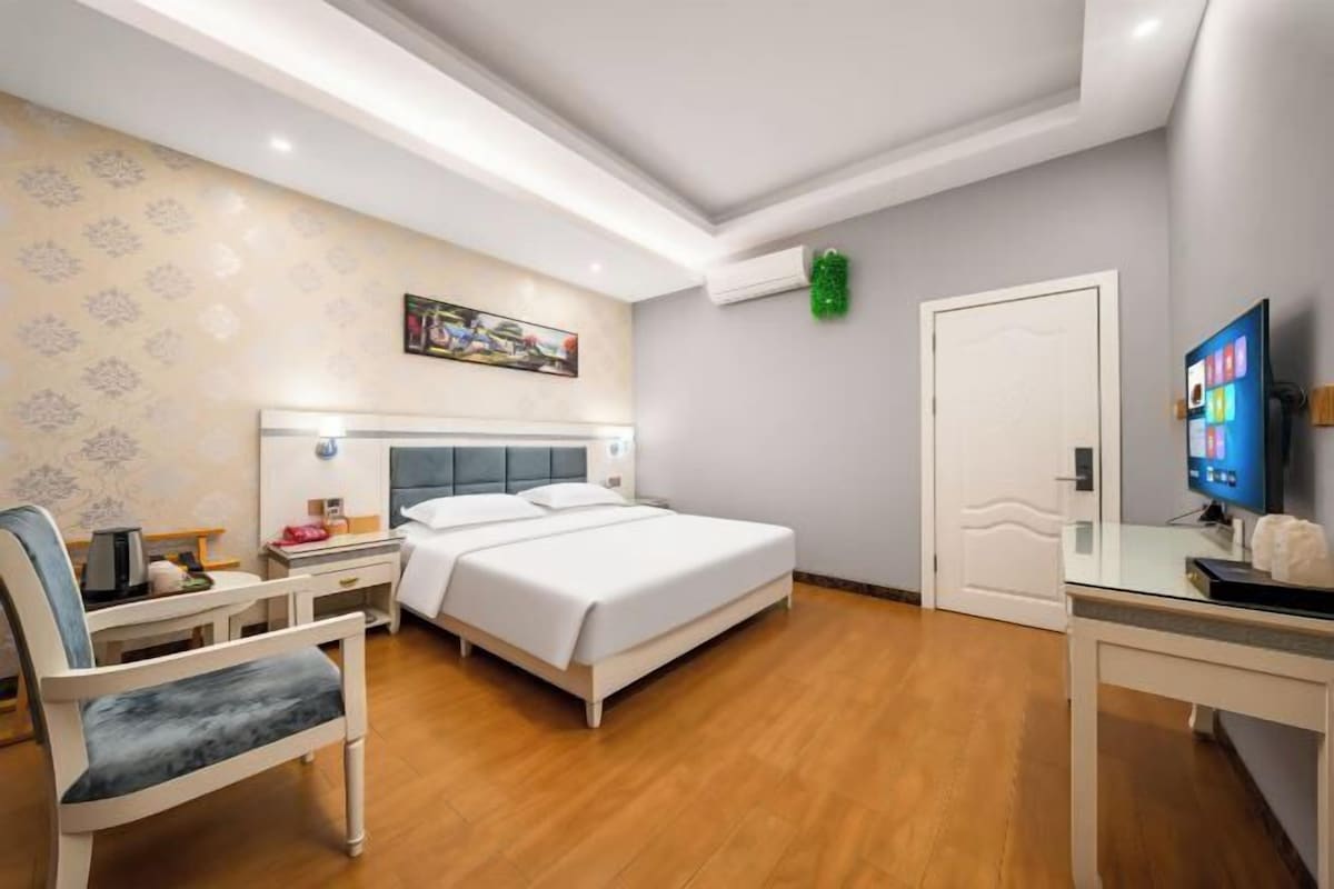 Habitación de hotel con cama, mesita de noche, silla, escritorio, televisor y puerta.