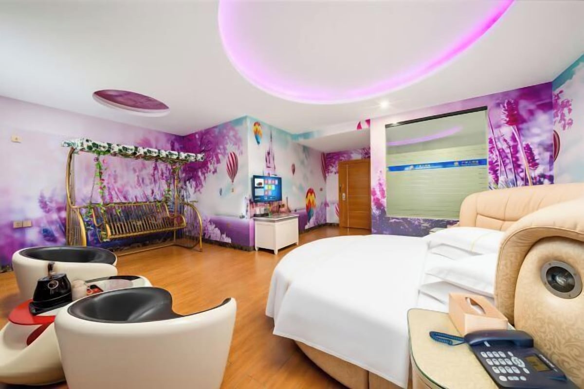 Habitación de hotel con cama, escritorio con computadora, silla, sillón de masaje y un mural colorido en la pared.