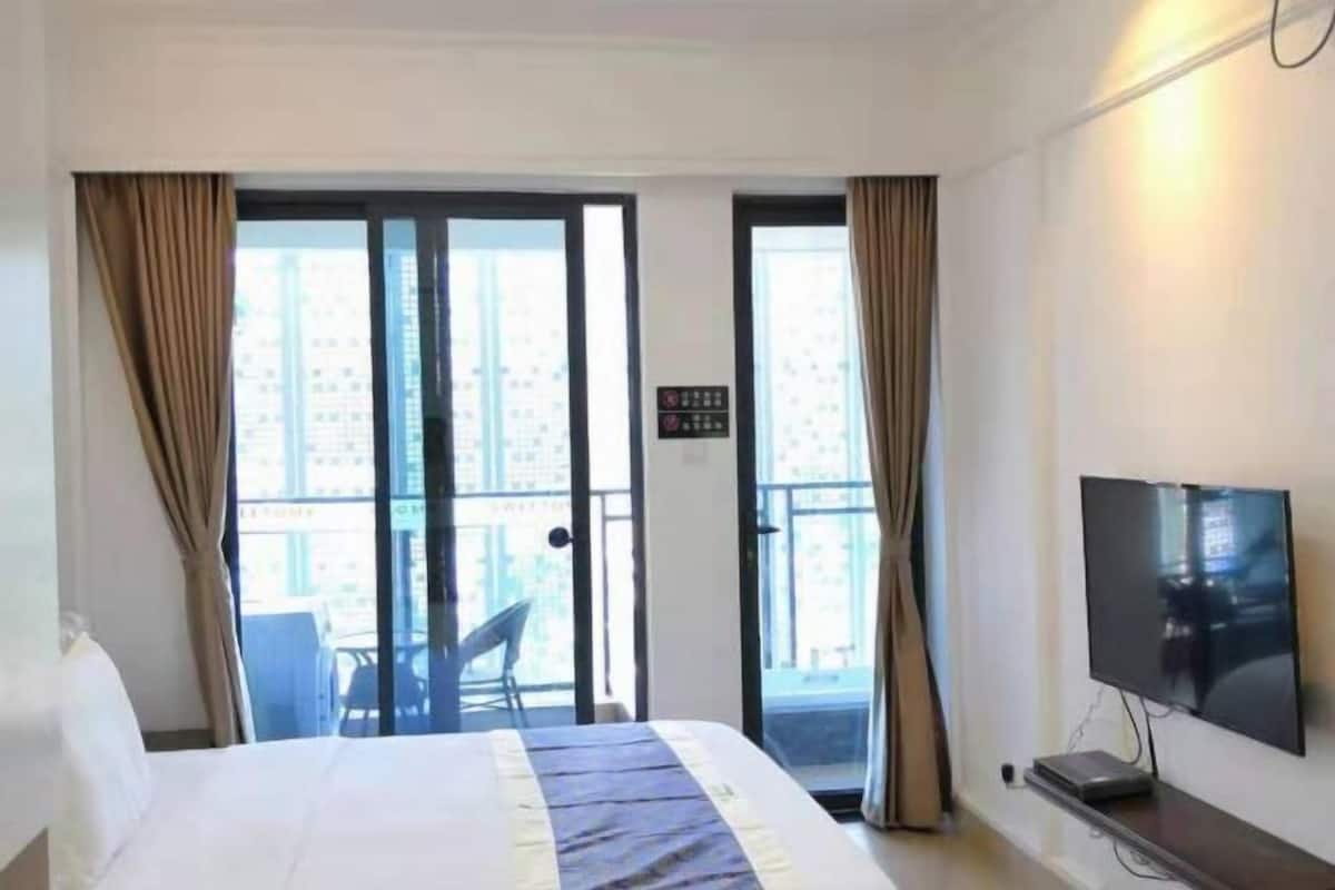 Habitación de hotel con cama, televisión, balcón con silla y vista a edificios.