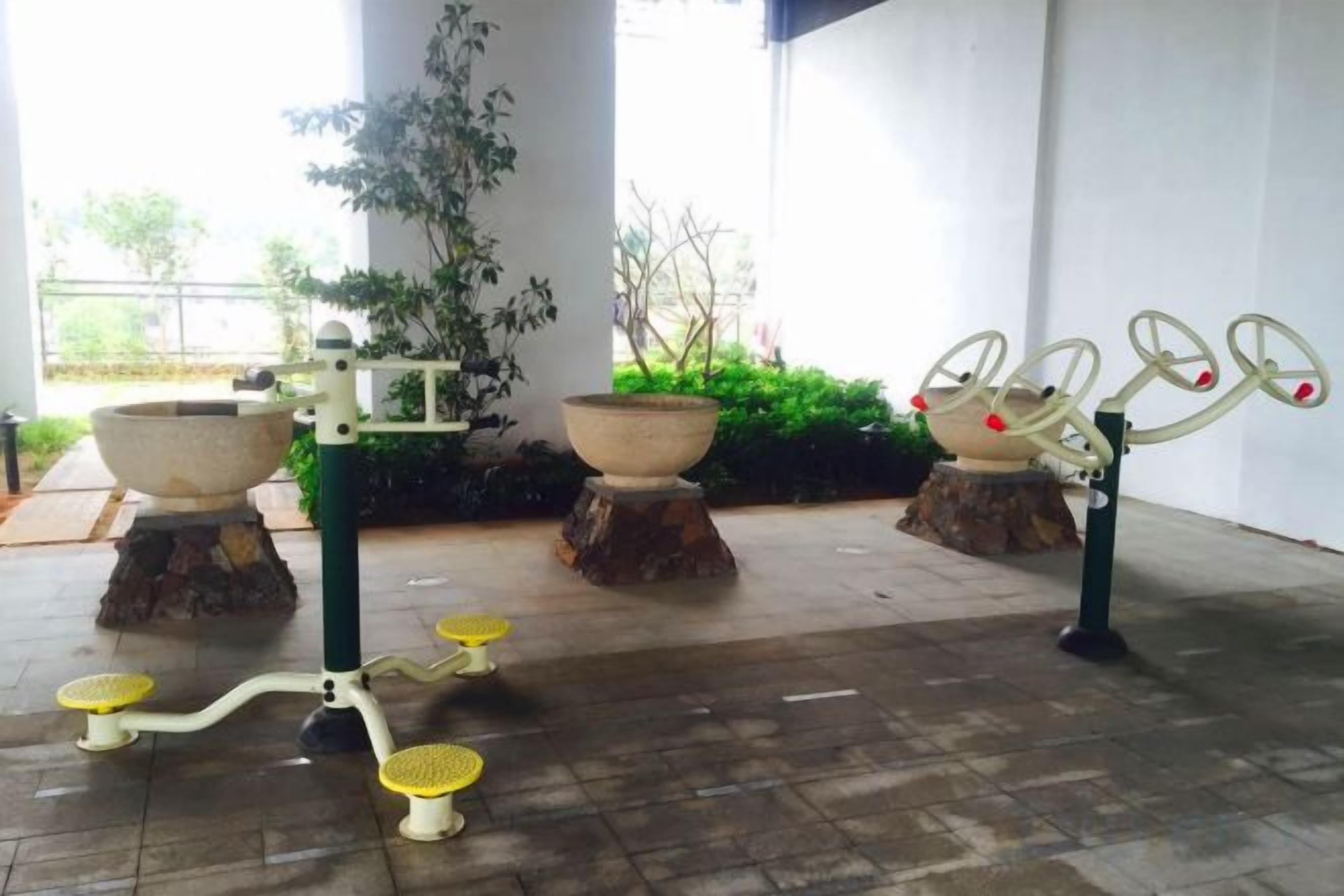 Buitenfitnessapparatuur, waaronder een hometrainer en een roeimachine, met op de achtergrond potplanten en een gebouw.