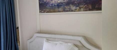 Una cama con cabecera, un almohadón blanco y un cuadro encima.