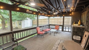Un patio cubierto con muebles de madera, luces colgantes y vistas a un jardín trasero.
