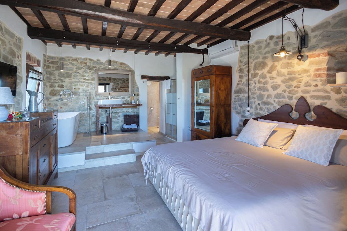 A bedroom with a large bed, a wooden wardrobe, a stone wall, and a bathroom with a sink and mirror.