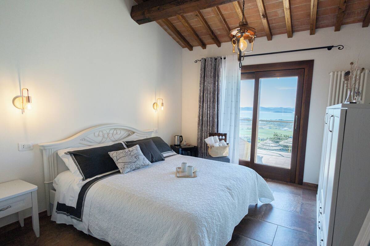 A bedroom with a bed, bedside tables, a nightstand, a lamp, a window with curtains, and a view of the outdoors.