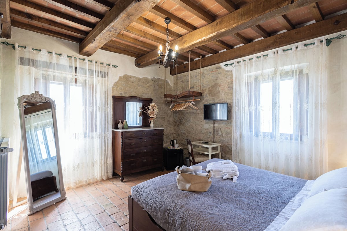 A bedroom with a large bed, wooden ceiling beams, a dresser, a mirror, a television, and a window with curtains.