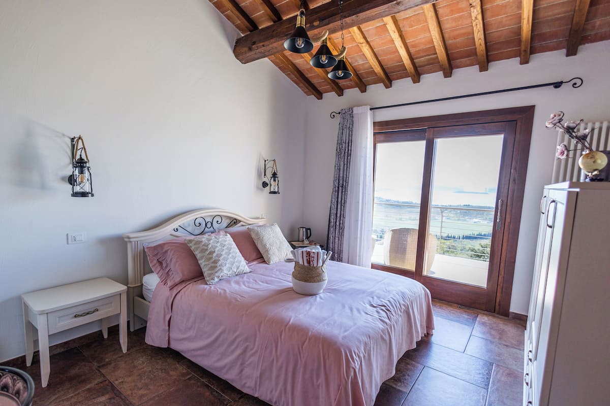 A bedroom with a bed, bedside table, lamp, and a view of the outdoors.