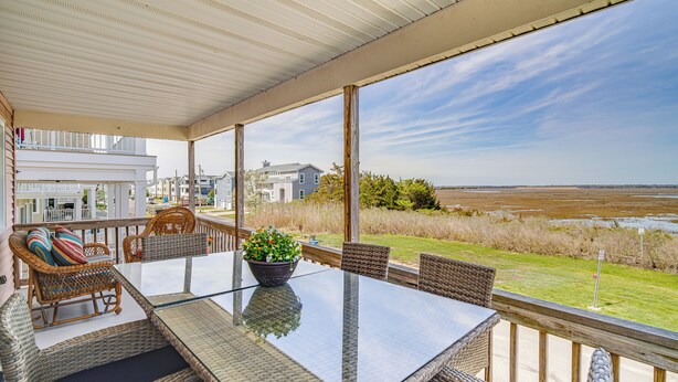 A covered outdoor dining area with wicker furniture and a glass table, overlooking a grassy landscape and a body of water. - Walk to Beach: Ocean City Home w/ Deck & Views! (Ocean City)