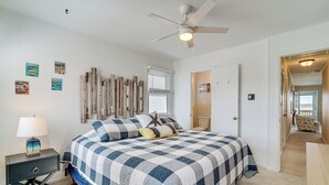 A bedroom with a large bed, a ceiling fan, a nightstand, and a wall with wooden panels and artwork. - Walk to Beach: Ocean City Home w/ Deck & Views! (Ocean City)