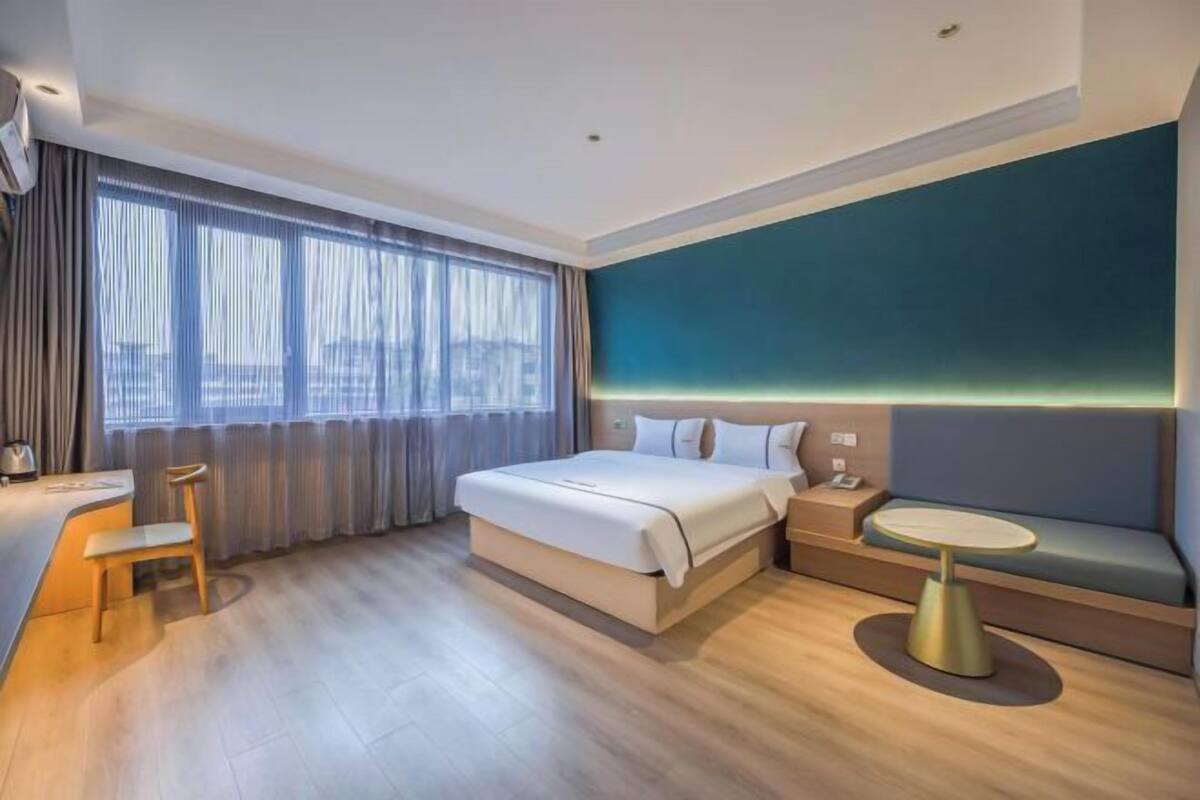 Habitación de hotel con cama, escritorio, silla, mesa redonda y vistas a la ciudad a través de amplios ventanales.