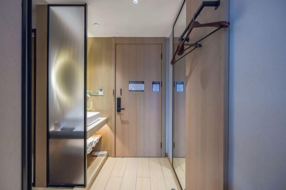 A modern hotel room with a wooden door, a small bathroom with a sink and mirror, and a wall-mounted towel rack.