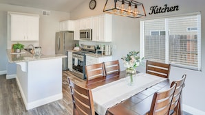 A modern kitchen with white cabinetry, a central island, and a dining area with wooden chairs. - Bakersfield Home w/ Pool Table, Cornhole + More! (Bakersfield)