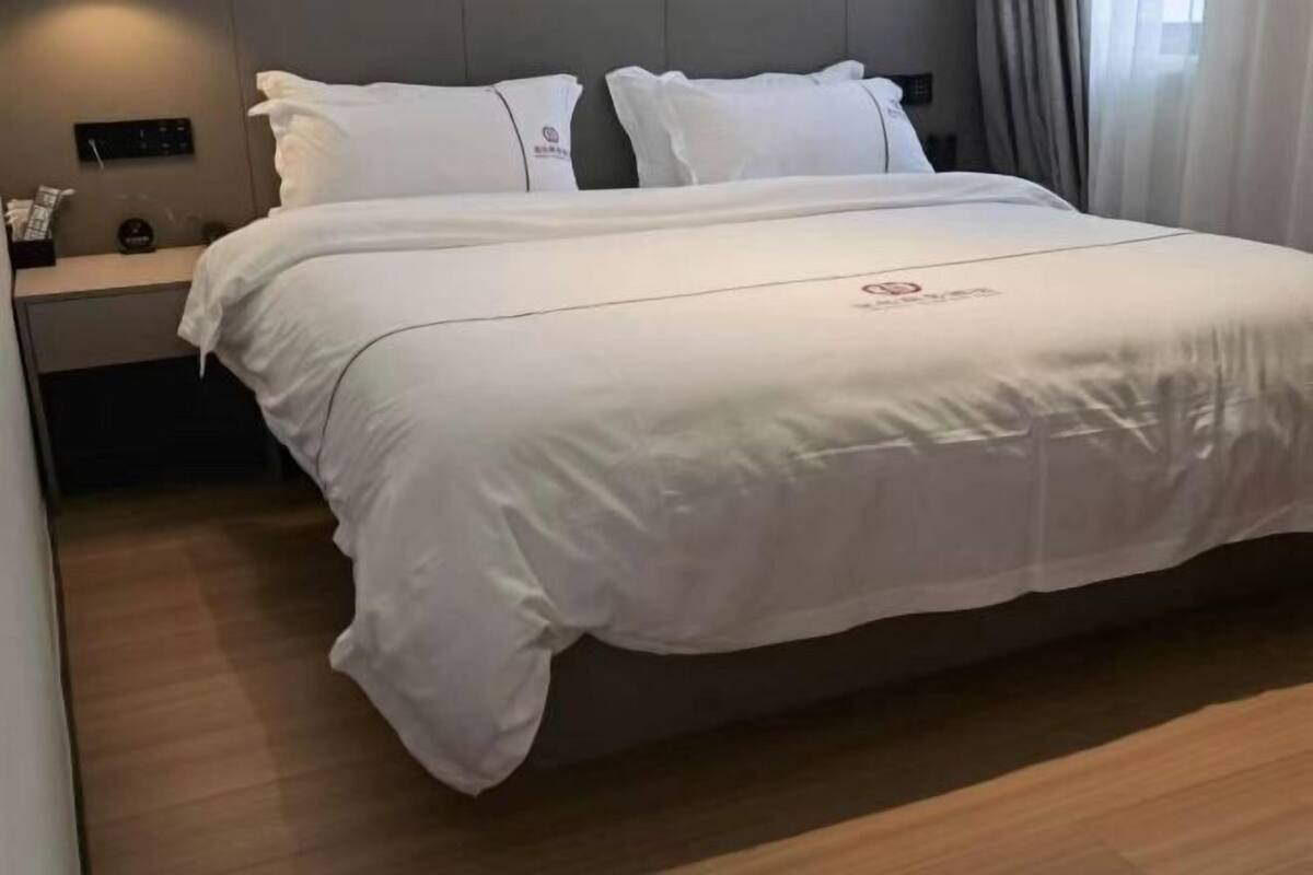 A neatly made bed with white bedding and pillows, a bedside table with a clock and a small decorative item, and a wooden floor.
