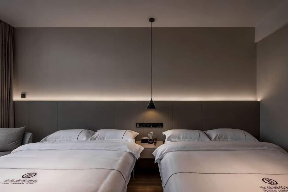 A hotel room with two beds, a bedside table with a lamp, and a wall-mounted control panel.