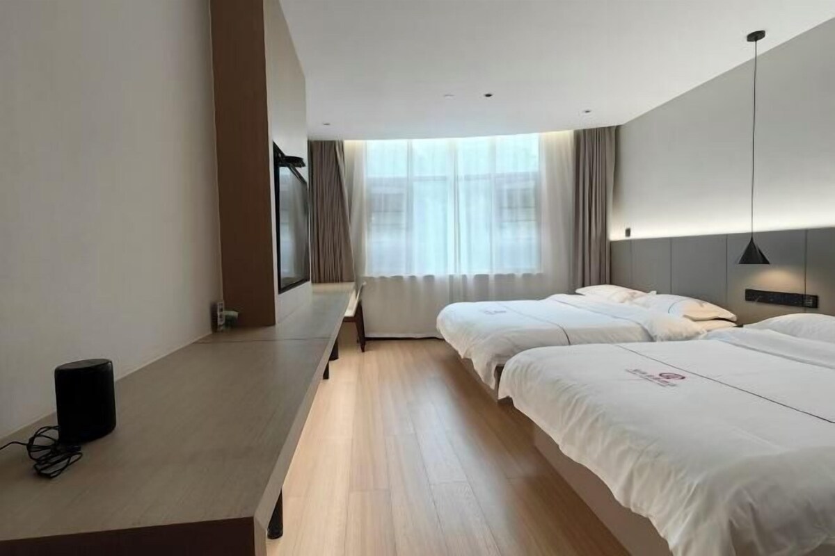 A hotel room with two beds, a desk with a speaker, and a large window with curtains.