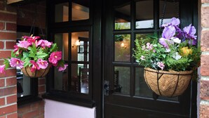 Dos cestas de flores colgando de una puerta negra, con un muro de ladrillo y ventanas al fondo.
