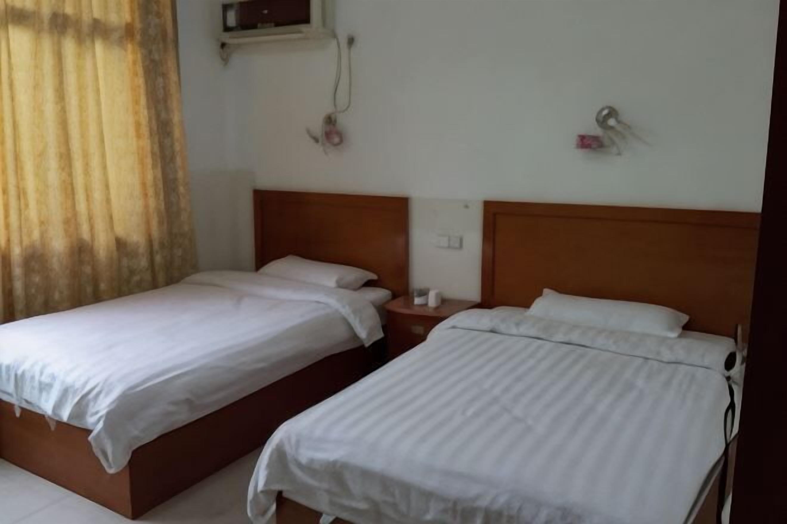 A hotel room with two single beds, a wooden headboard, a bedside table, and a wall-mounted air conditioner.