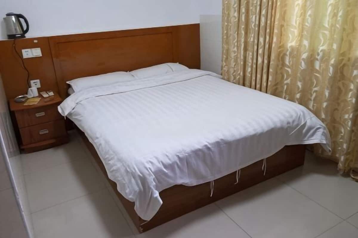 Una cama bien tendida con ropa de cama blanca, un cabecero de madera, una mesita de noche con lámpara y una cortina.