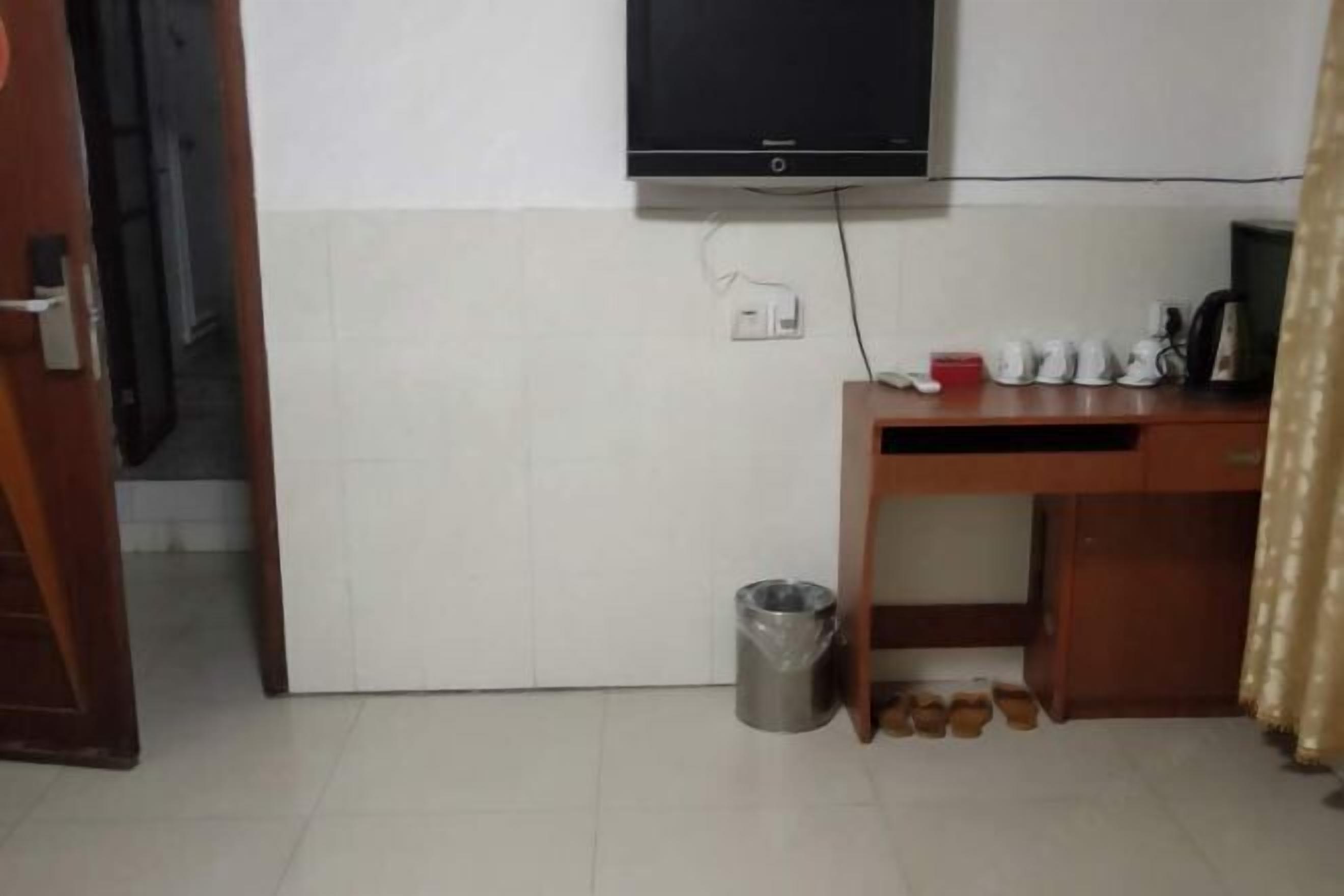 Ein Zimmer mit an der Wand montiertem Fernseher, einem Schreibtisch mit Computer, einem Mülleimer und einem Schuhregal.