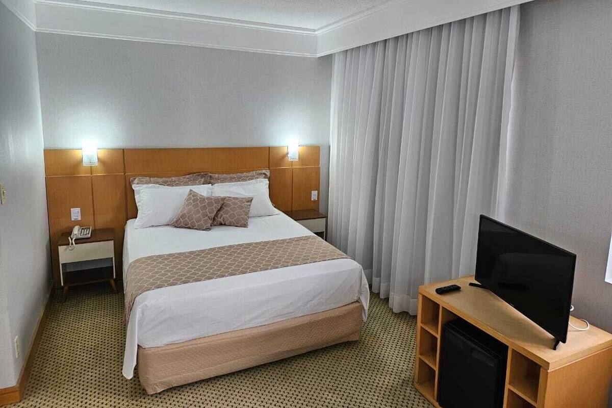 A hotel room with a bed, bedside lamps, a television on a wooden stand, and a curtain on the window.