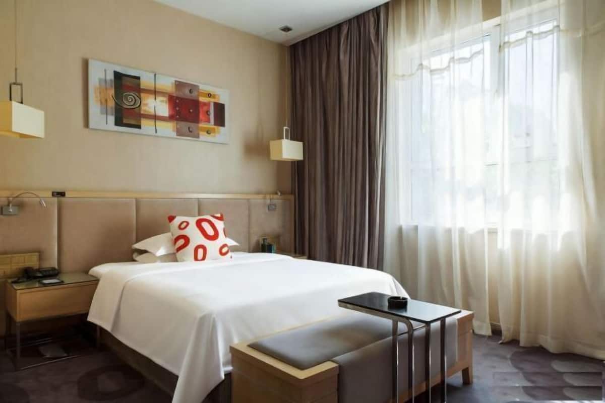 A hotel room with a bed, bedside tables, a small table, a bench, a painting, and a window with curtains.