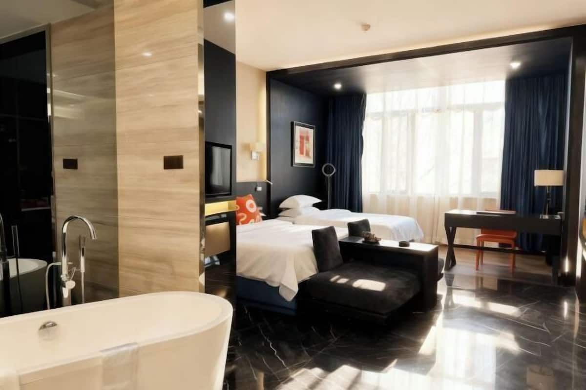 A modern hotel room with a large bathtub, a bed with two pillows, a desk, and a large window with curtains.