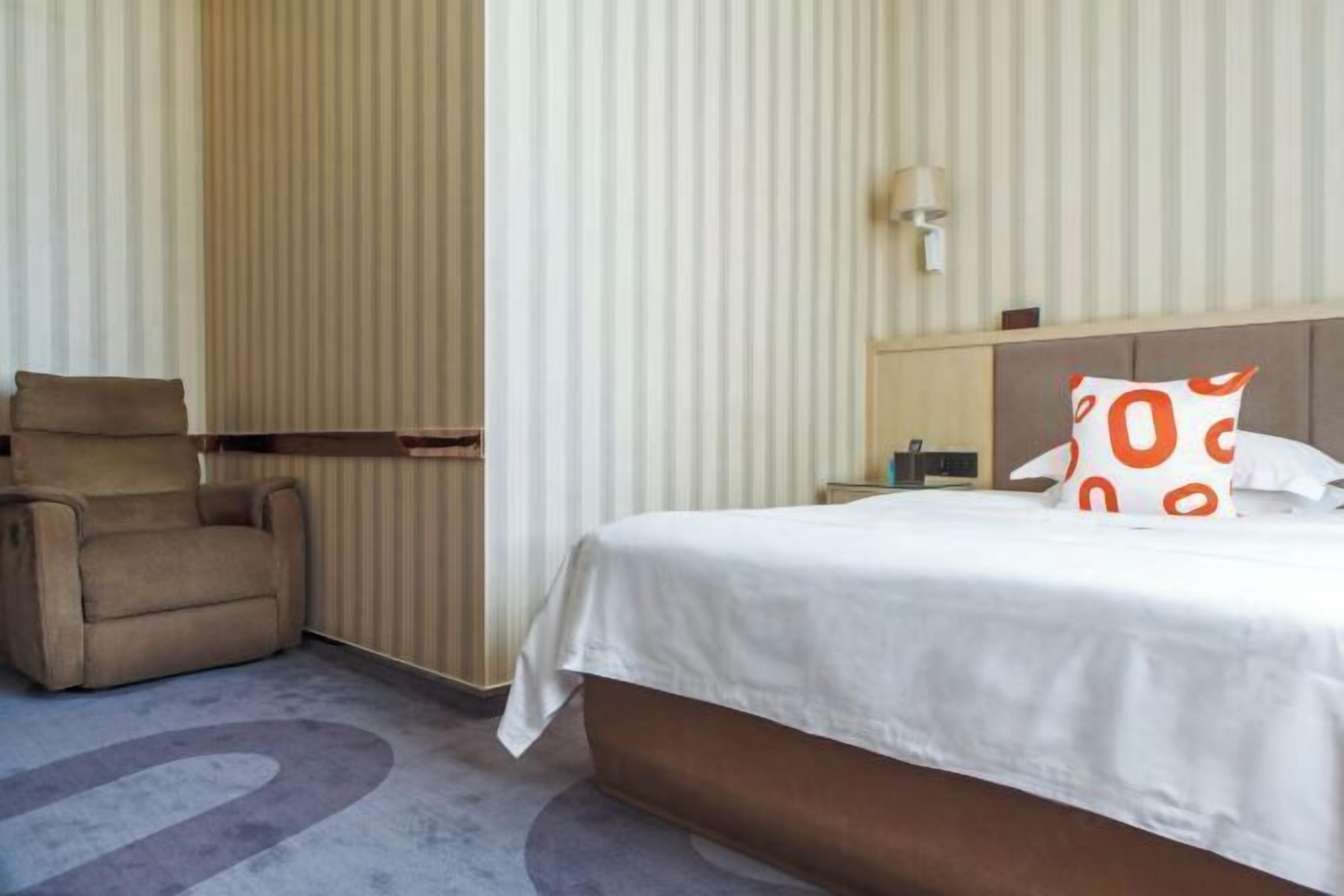 A hotel room with a bed, a chair, a nightstand, and a wall-mounted lamp.