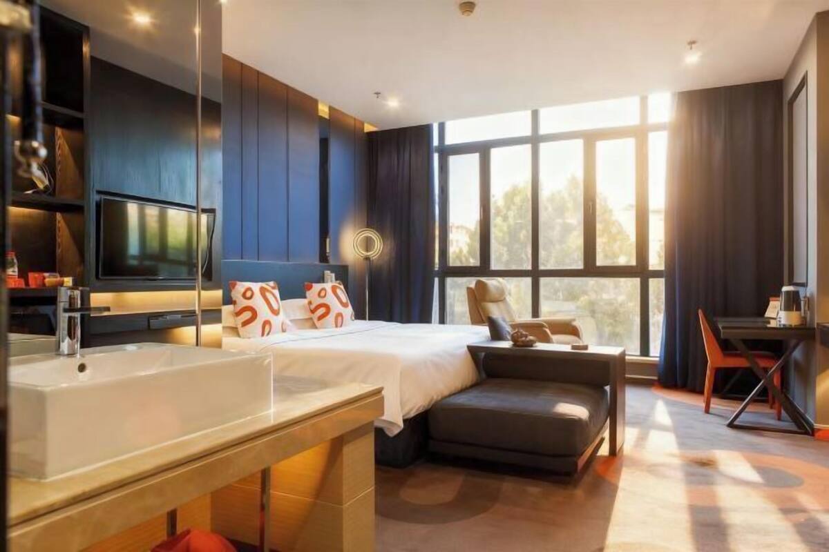 A modern hotel room with a large bed, a desk, a sofa, a TV, and a large window.