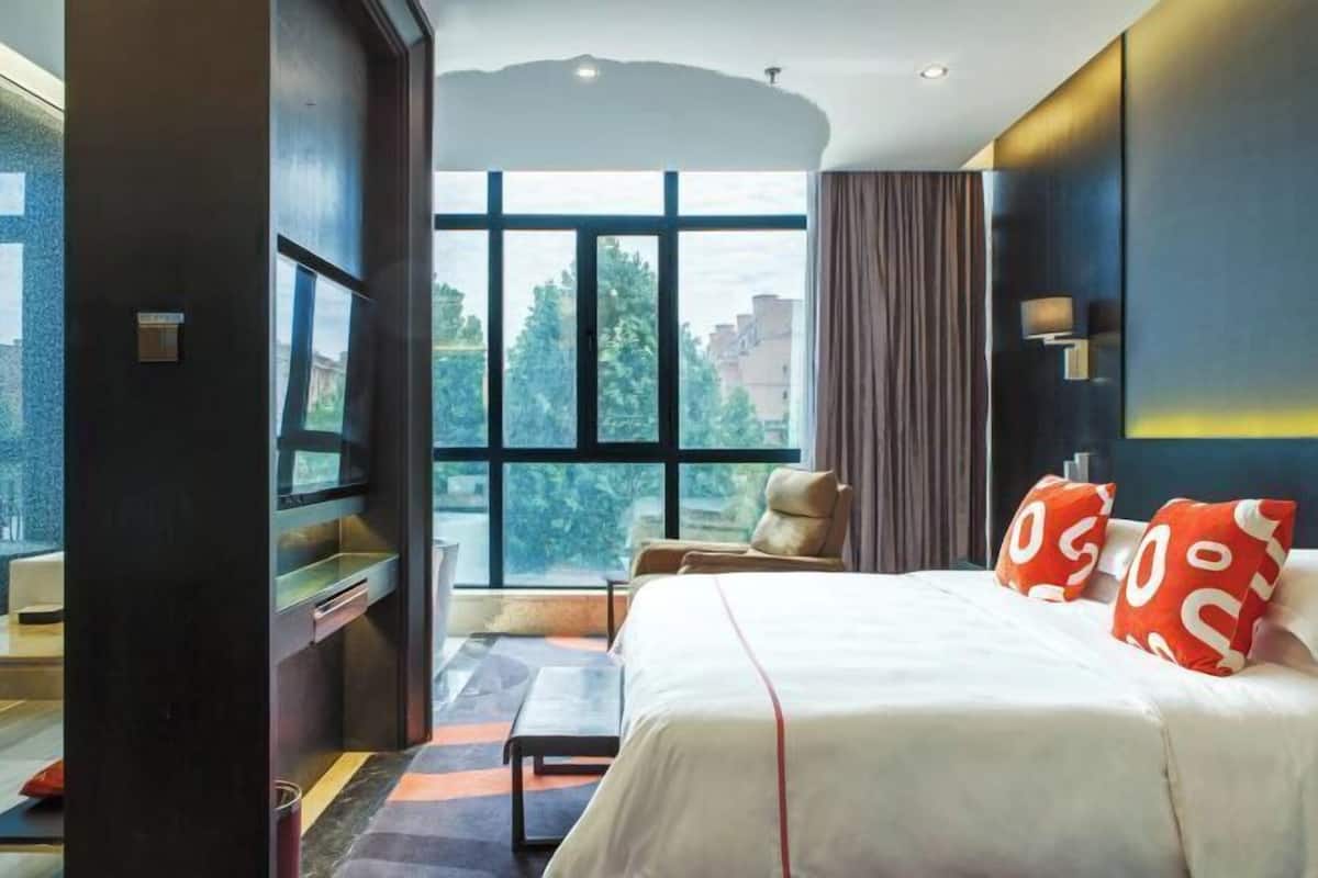 A hotel room with a large window, a bed with white bedding and red pillows, a chair, and a small table.