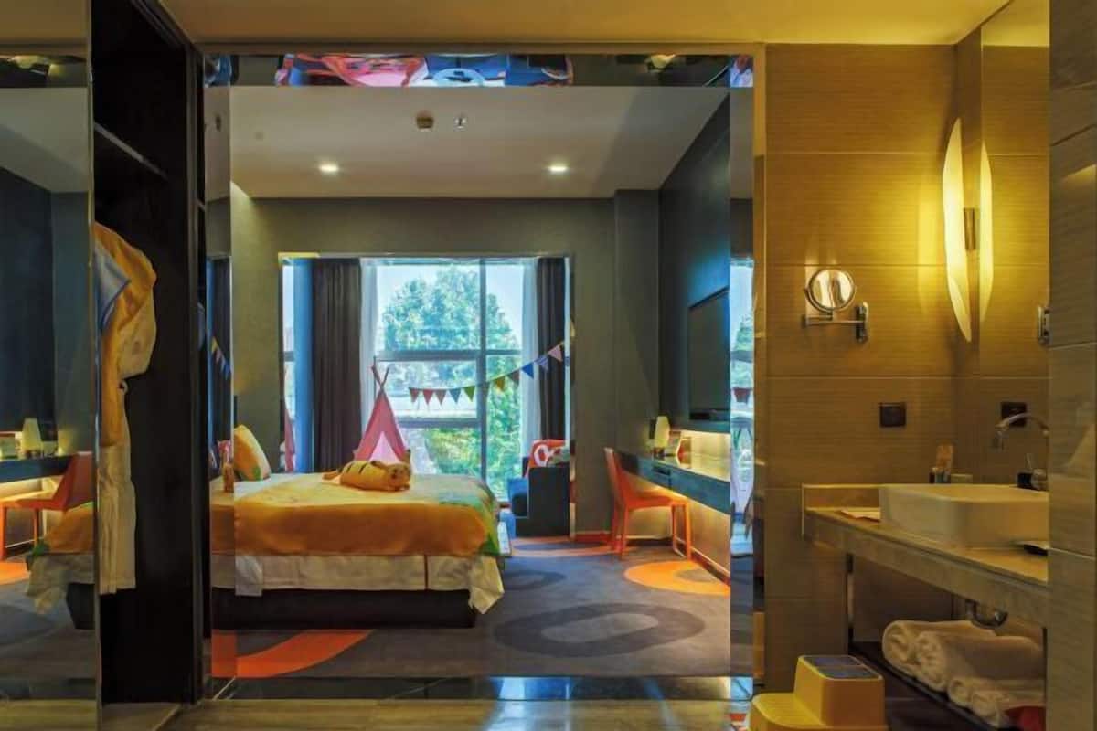 A hotel room with a bed, a desk, a chair, a window with curtains, a bathroom with a sink and mirror, and a view of trees outside.
