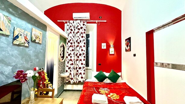 Una habitación de hotel compacta con una pared de acento rojo, una cama con almohadas verdes, una pequeña mesa con un jarrón de flores y una zona de ducha con cortina estampada.
