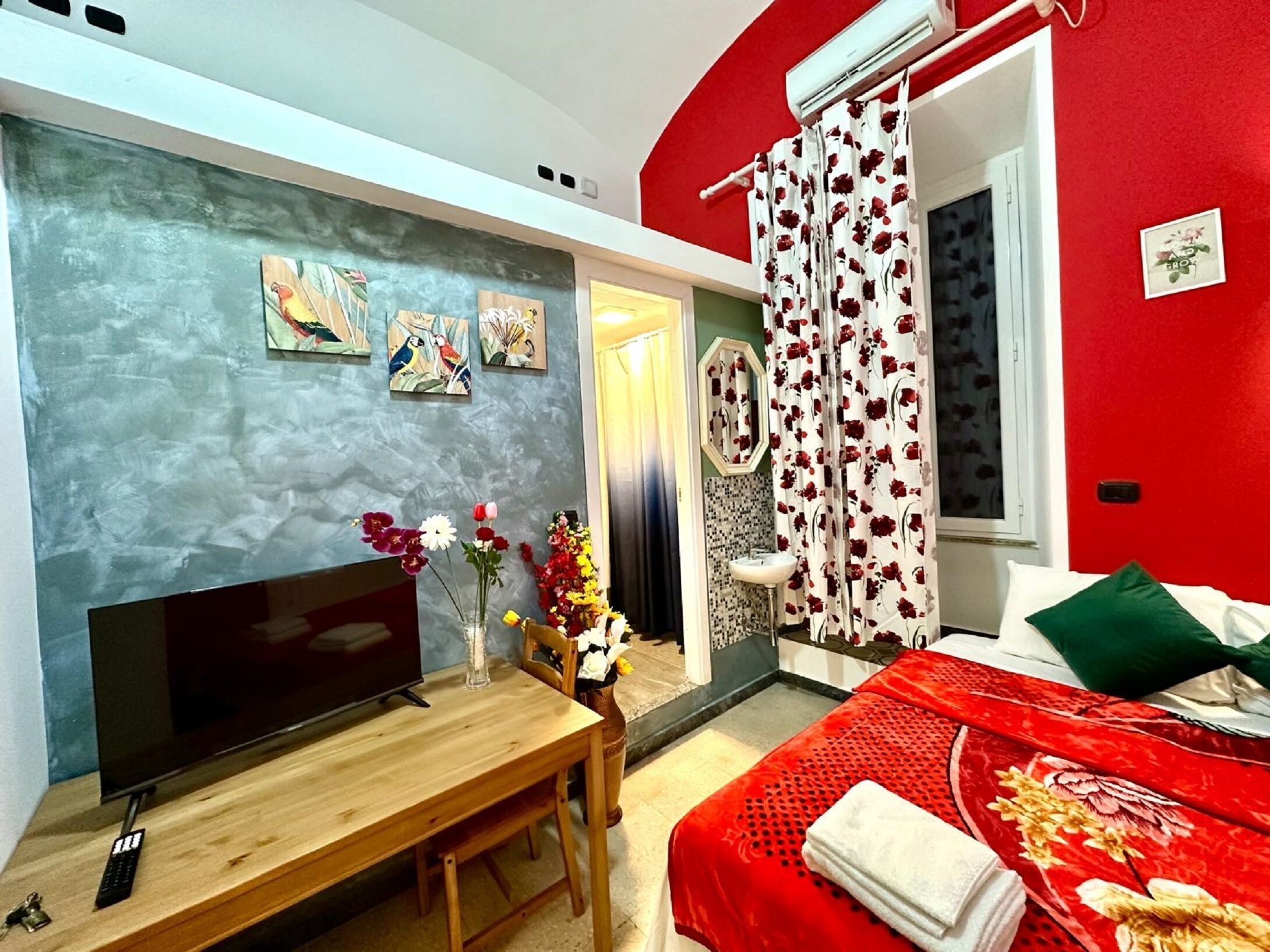 A bedroom with a TV, a wooden table, a bed with a red bedspread, a mirror, and a window with red curtains.