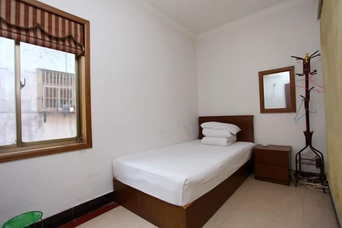 A small, clean room with a single bed, a nightstand, a mirror, and a window with a view of the outside.