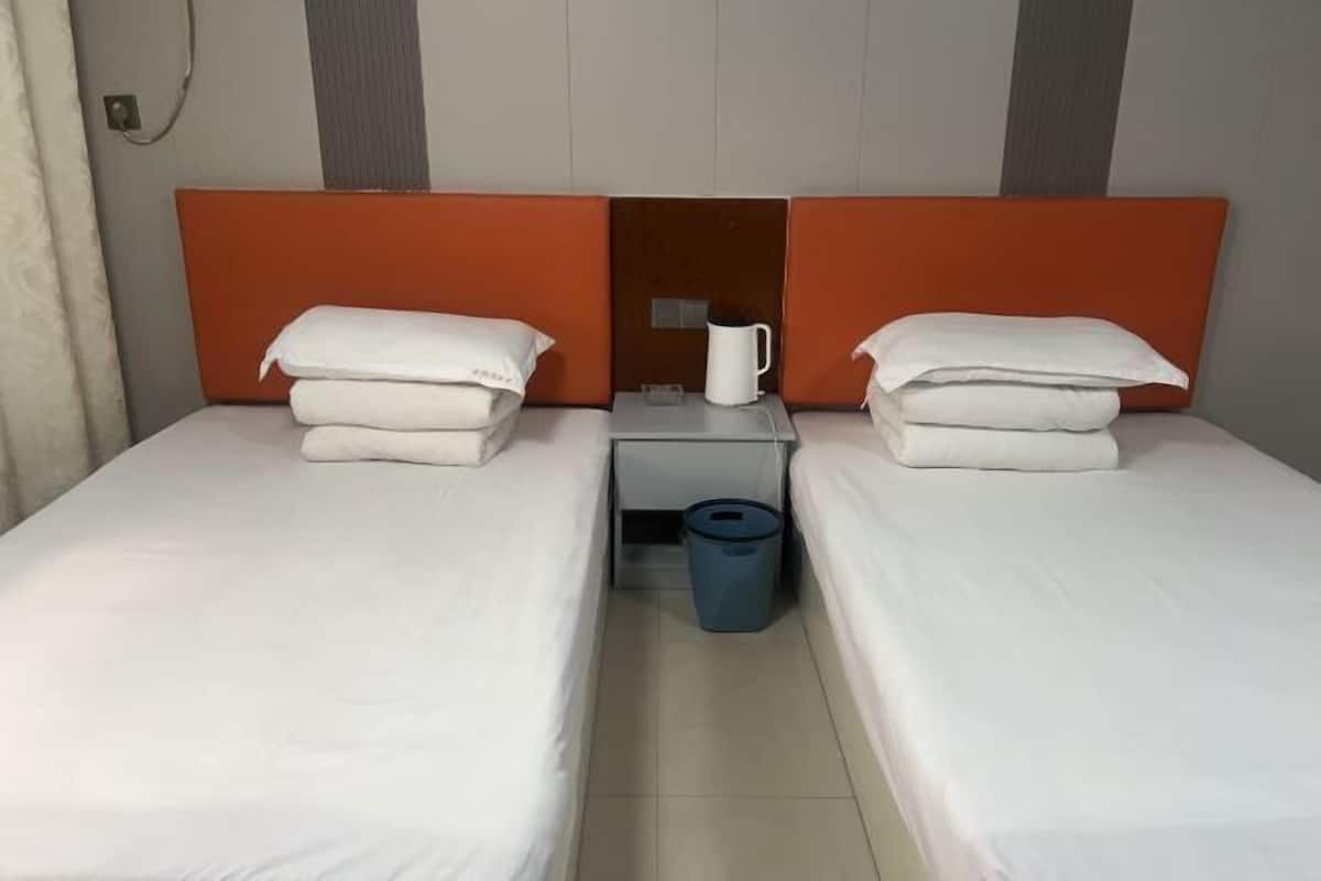 A hotel room with two single beds, each with white linens and pillows, a small table with a white mug and a trash bin, and an orange headboard.