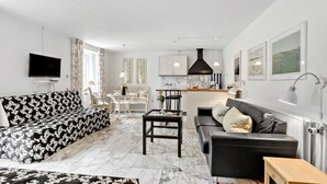 Ein modernes Wohnzimmer mit einem schwarz-weiß gemusterten Sofa, einem schwarzen Ledersessel und einem Boden mit Marmorplatten. Es gibt einen Essbereich mit weißem Tisch und Stühlen sowie eine Küche mit weißen Schränken und einer schwarzen Dunstabzugshaube.