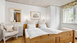 Ein Schlafzimmer mit einem hölzernen Bett, einem Korbstuhl, einem Nachttisch, einem Kleiderschrank und einem Fenster mit Vorhängen.